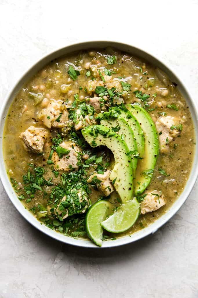 Sliced avocados placed on top of a bowl of green chicken chili with lime wedges on the side. 