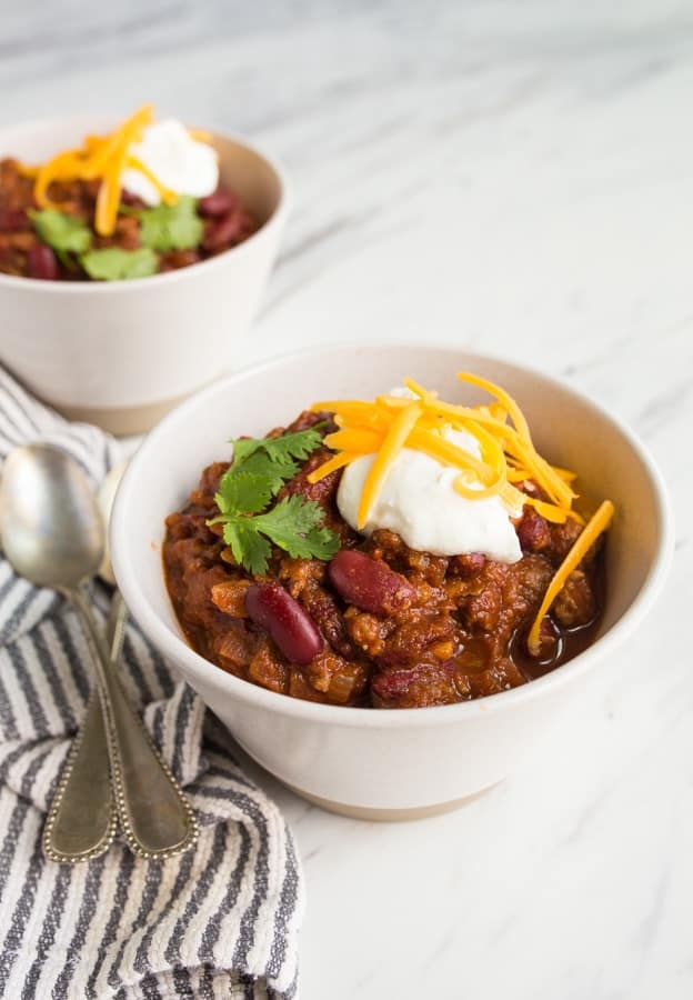 Two spoons sitting next to a bowl of chili with sour cream and shredded cheese garnished on otp. 