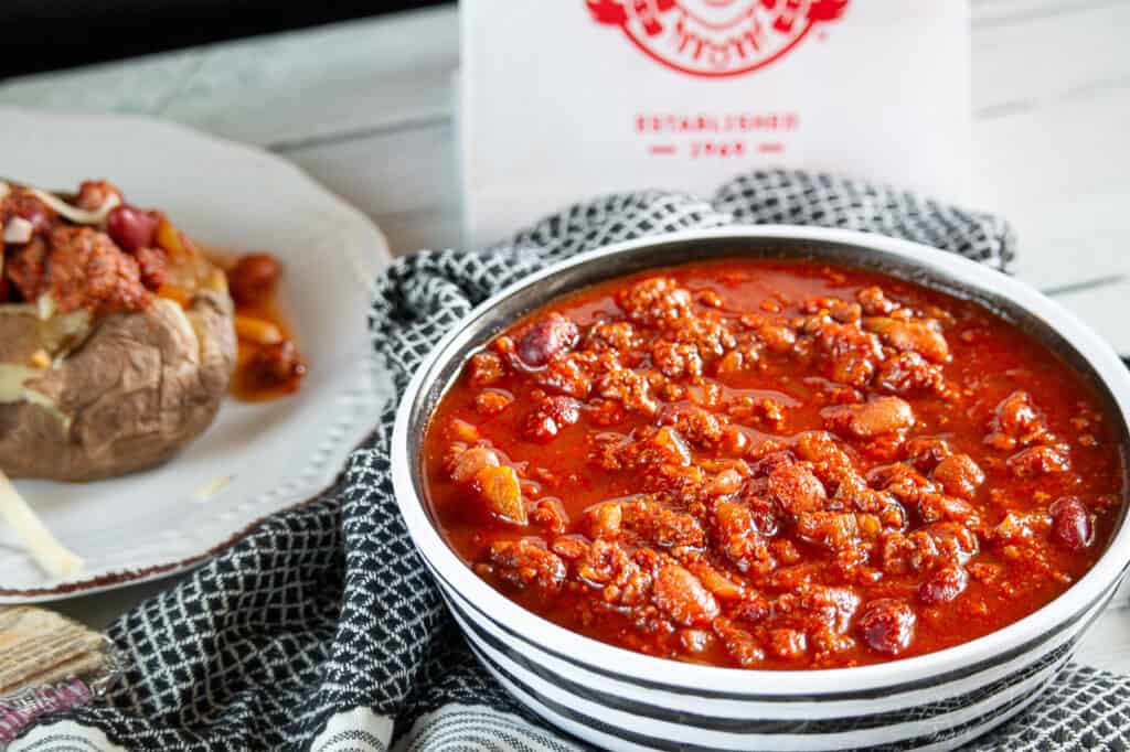 Bowl of homemade wendy's chili with a baked potato on a plate next to it with chili covering it. 