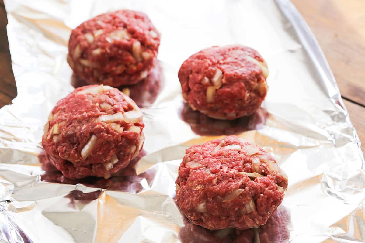 Four hamburger patties on a piece of foil, with onions mixed into the beef.