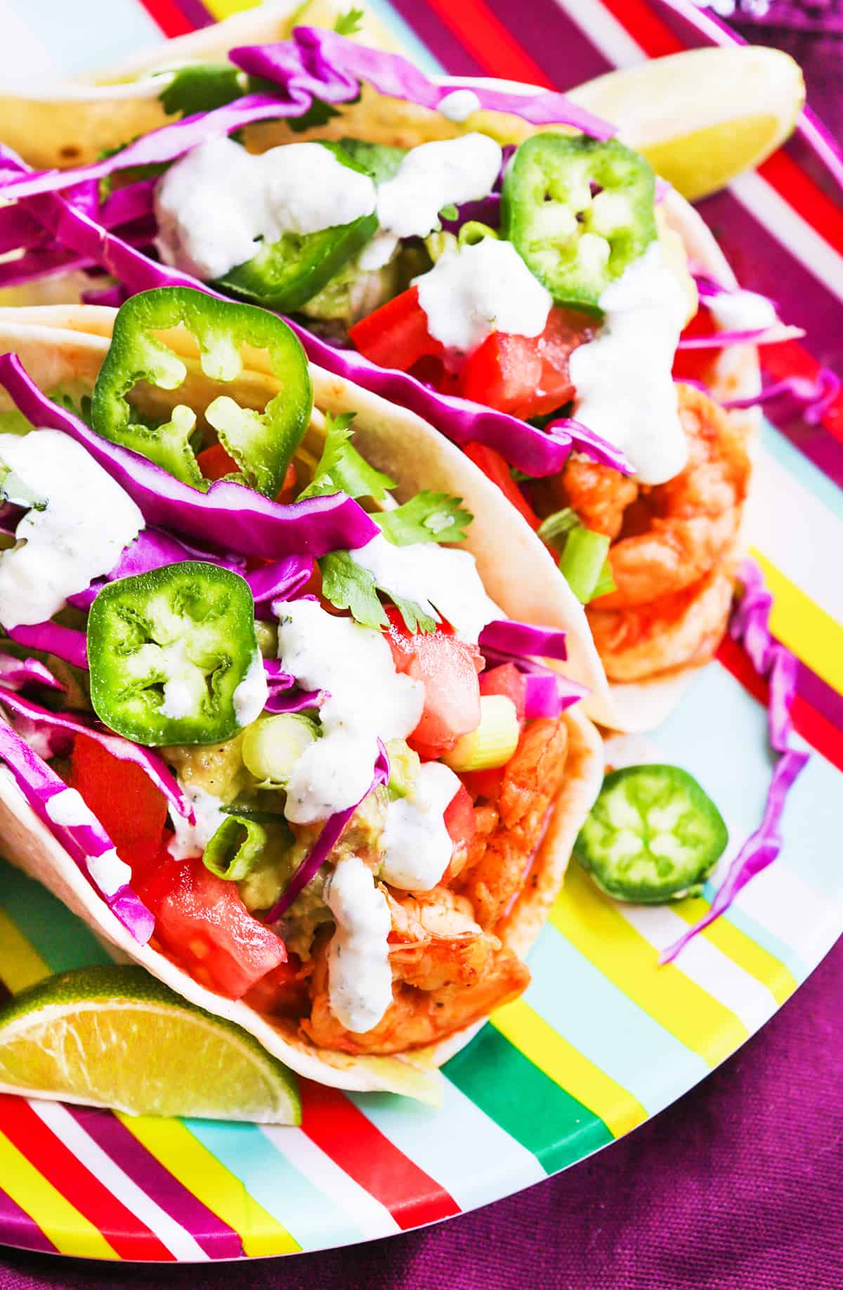Two shrimp tacos on a colorful plate, topped with cabbage, jalapeno slices and sauce.