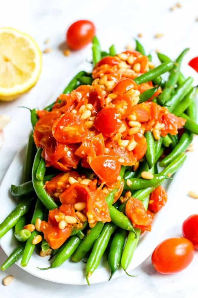 Oval serving platter filled with cooked green beans with pine nuts and grape tomatoes on top. 