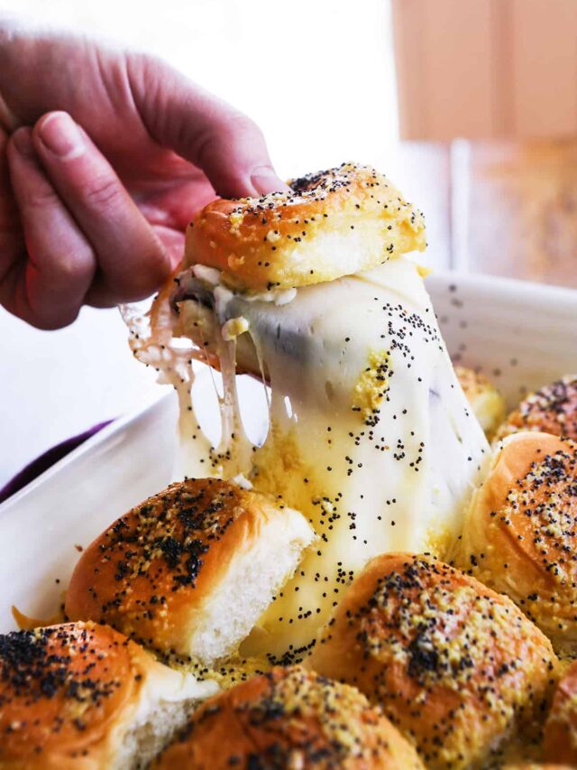 Hand picking up a ham and cheese slider from a full pan with gooey cheese pulling away from it. 