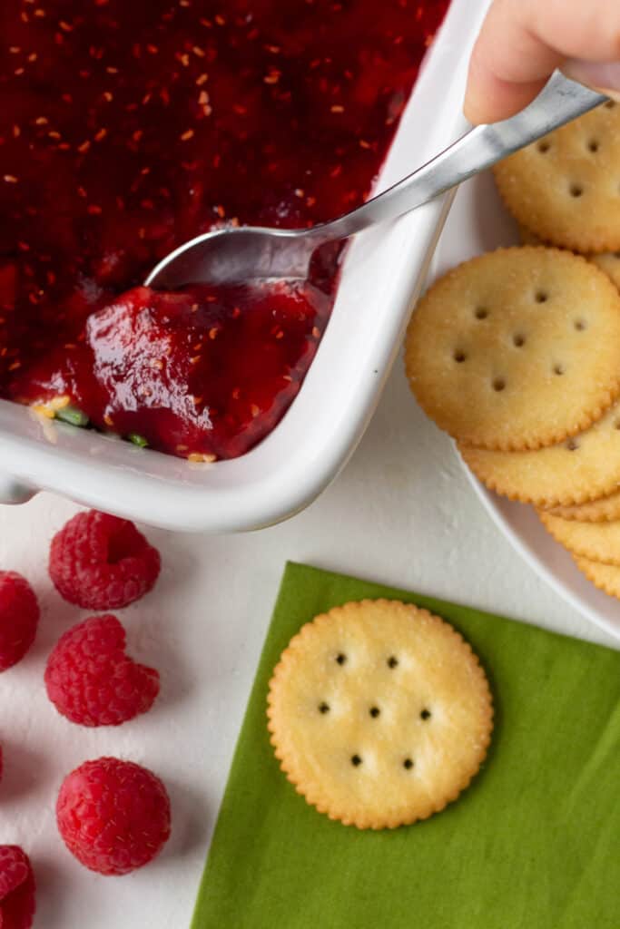 Spoon taking out a serving of raspberry dip with ritz crackers laying around the serving dish and fresh raspberries laying next to them. 