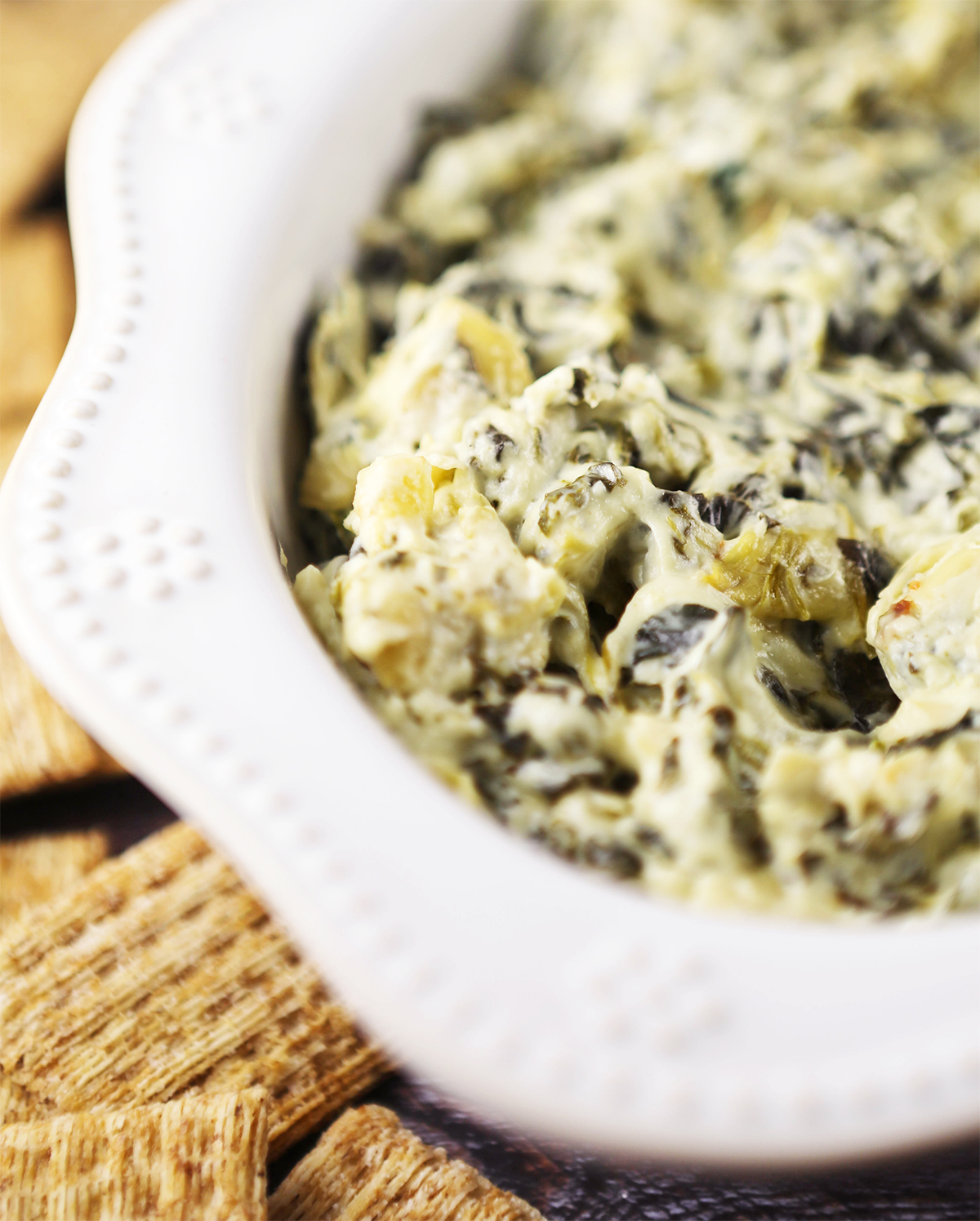 Closeup of a bowl filled with spinach artichoke dip, surrounded by Triscuit crackers.