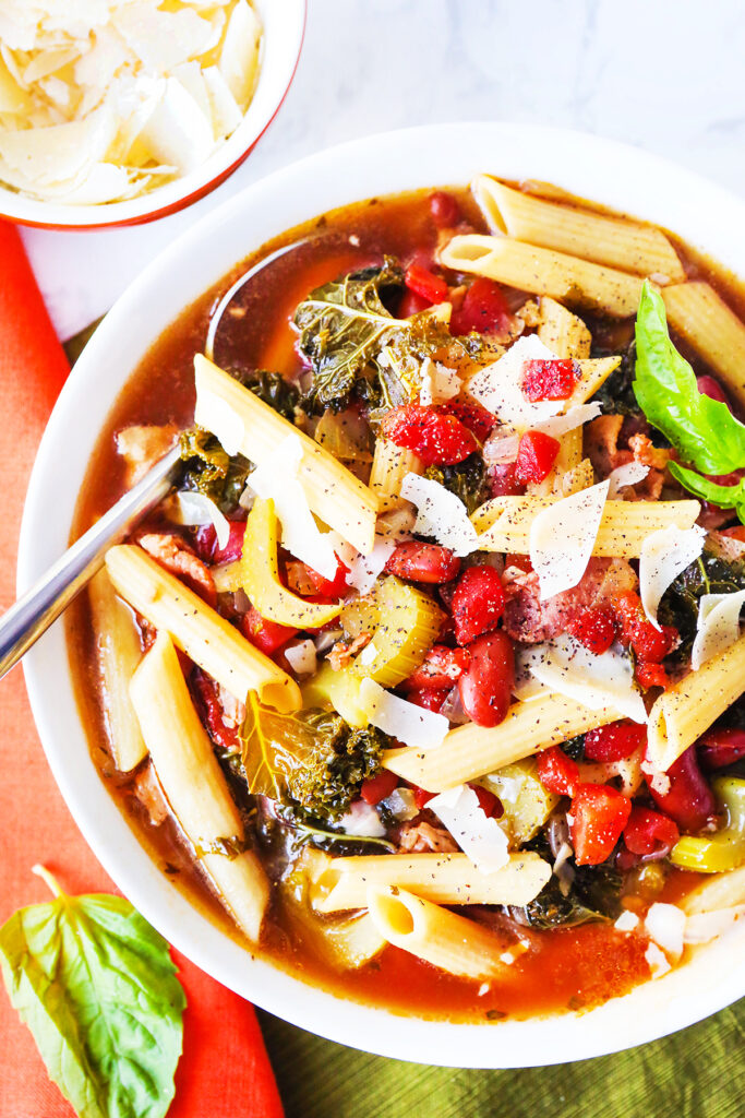 Top view of a bowl of Minestrone with pasta and shavings of parmesan on the top and a spoon tucked into the bowl. 