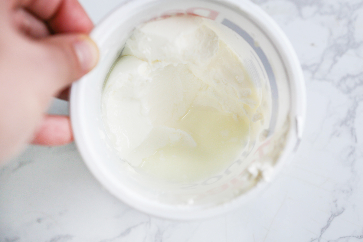 Hand holding a tub of sour cream with a bit of liquid inside.