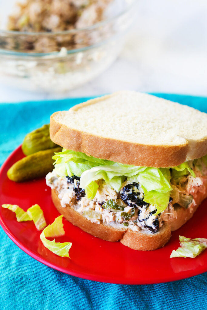 Chicken salad on white bread sitting on a red plate with baby dill pickles on the side. 