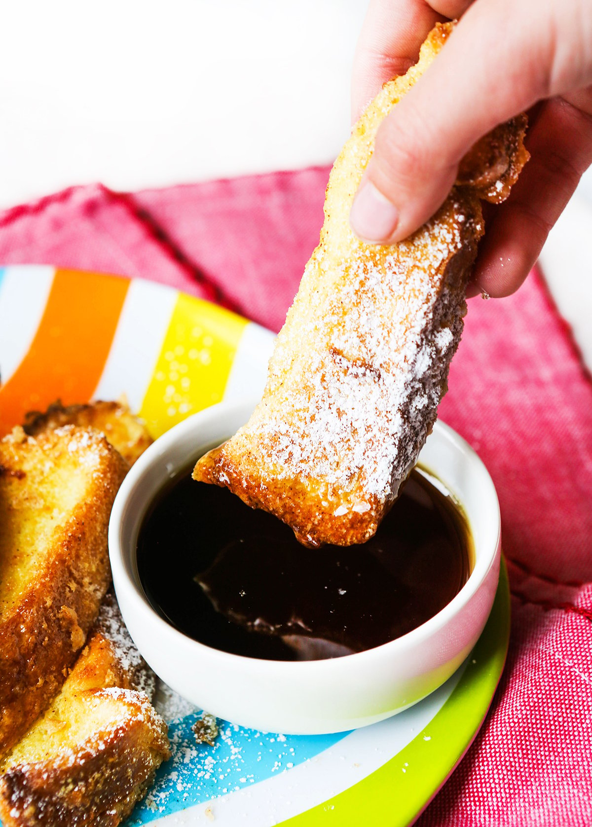 Hand dunking a French toast stick into a small bowl of maple syrup.