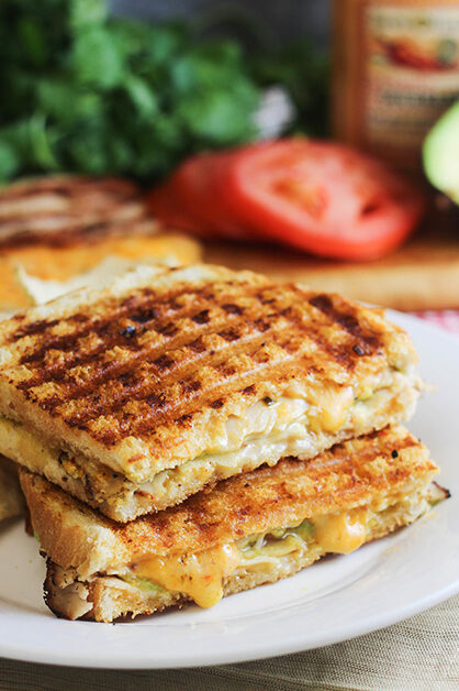 Two halves of a spicy southwestern turkey panini on a plate with fresh sliced tomatoes sitting behind it. 