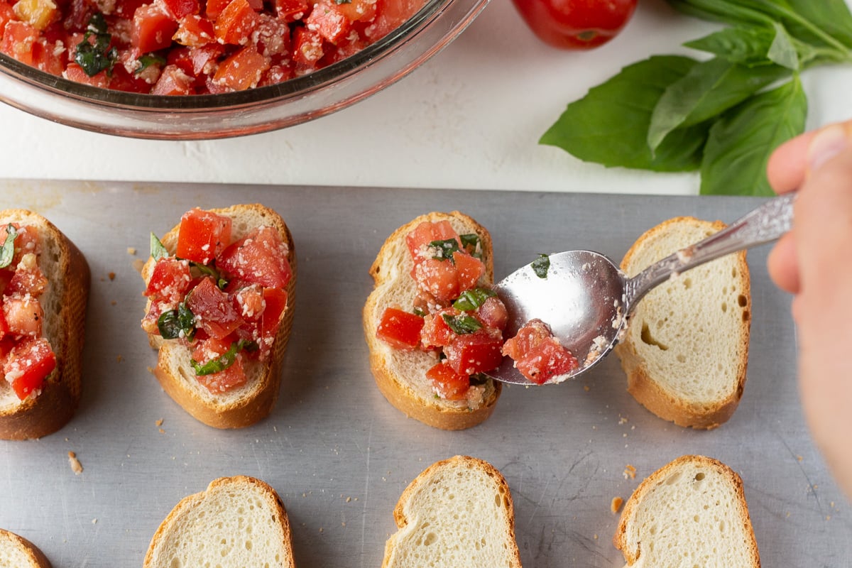 Bread slices on a baking sheet lined up, and a spoon dropping a tomato mixture over the top of a few of them.