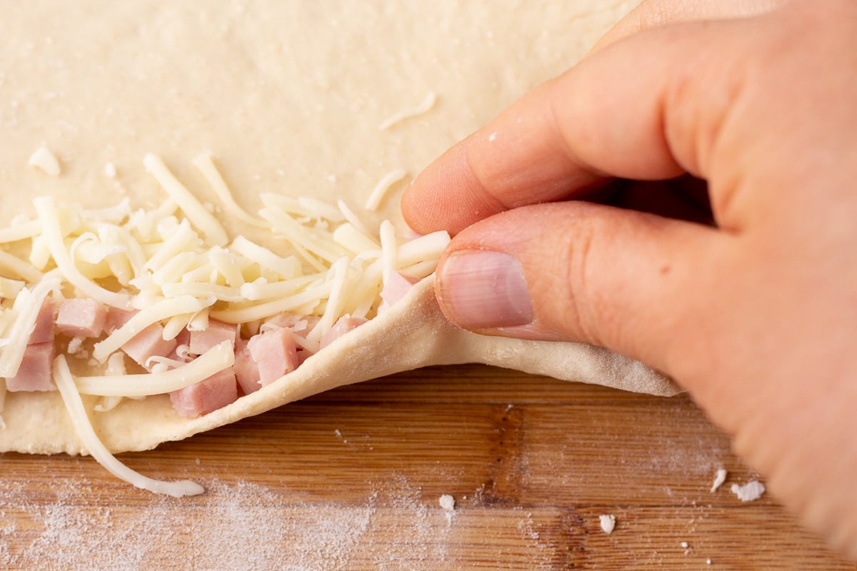Fingers pulling a long edge of dough over chopped ham and shredded cheese.