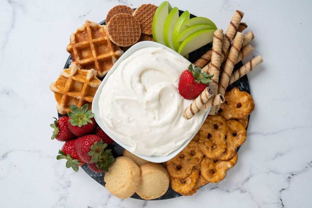 Cheesecake dip in a bowl with different treats to dip into it and munch on like fresh strawberries, apple slices, waffle bites and pretzel chips. 