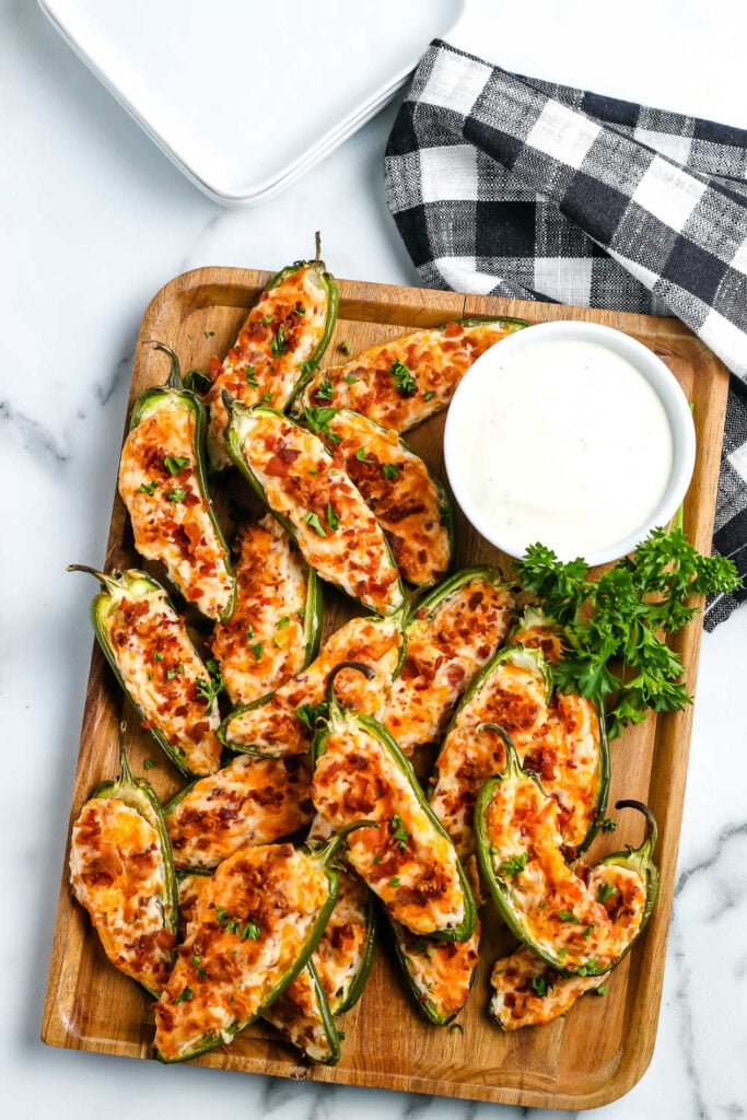 Baked jalapeno poppers on a cutting board with parsley as a garnish and dip in a bowl. 