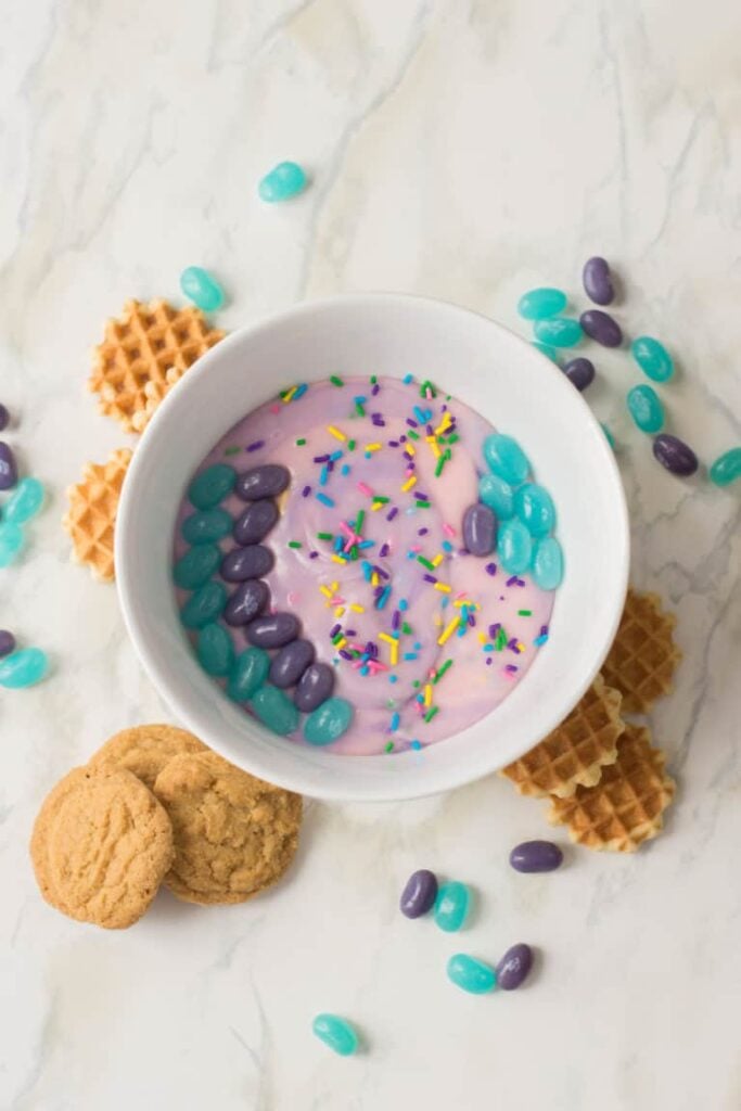 Mermaid cookie dip in a bowl with purple and blue jelly beans garnishing it and waffle cookies around the bowl. 
