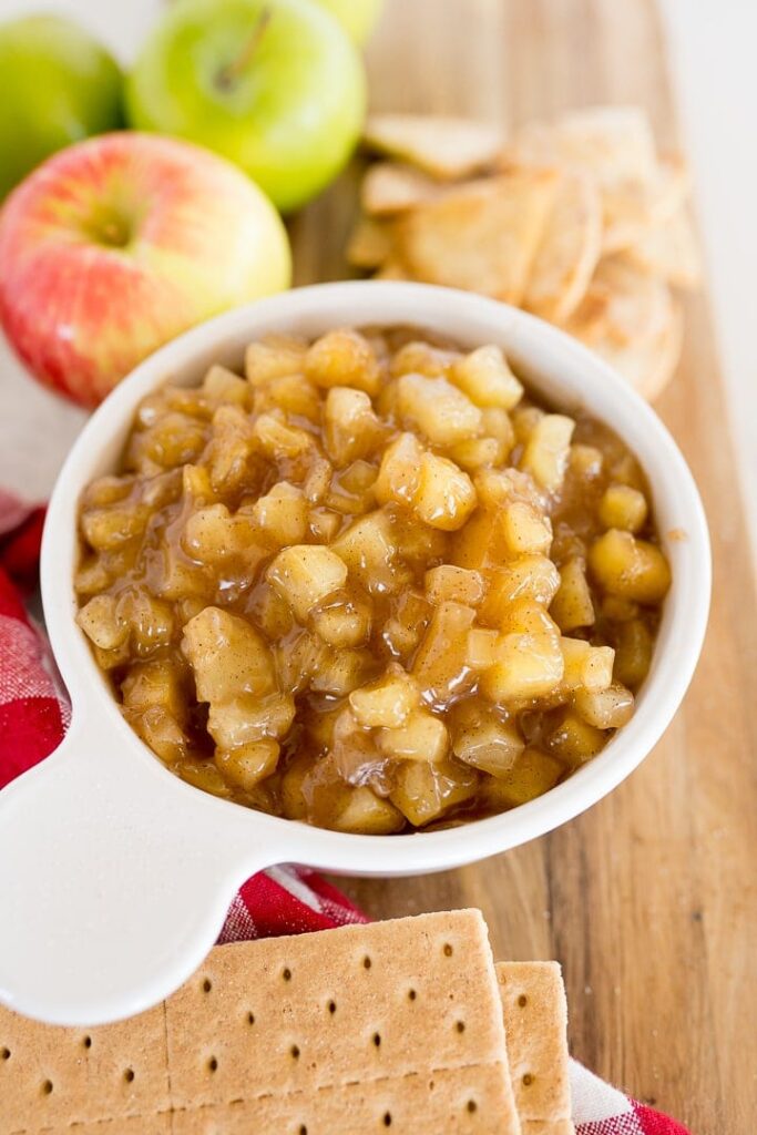 Whole apples sitting behind a crock of apple pie dip with graham crackers in front of it. 