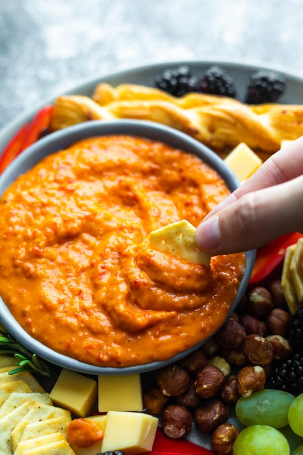 Hand using a cracker to scoop out red pepper dip. 