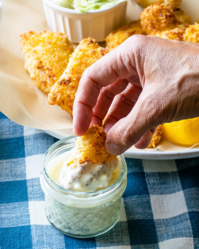 Hand dunking a fish piece into a small glass jar of homemade tartar sauce. 