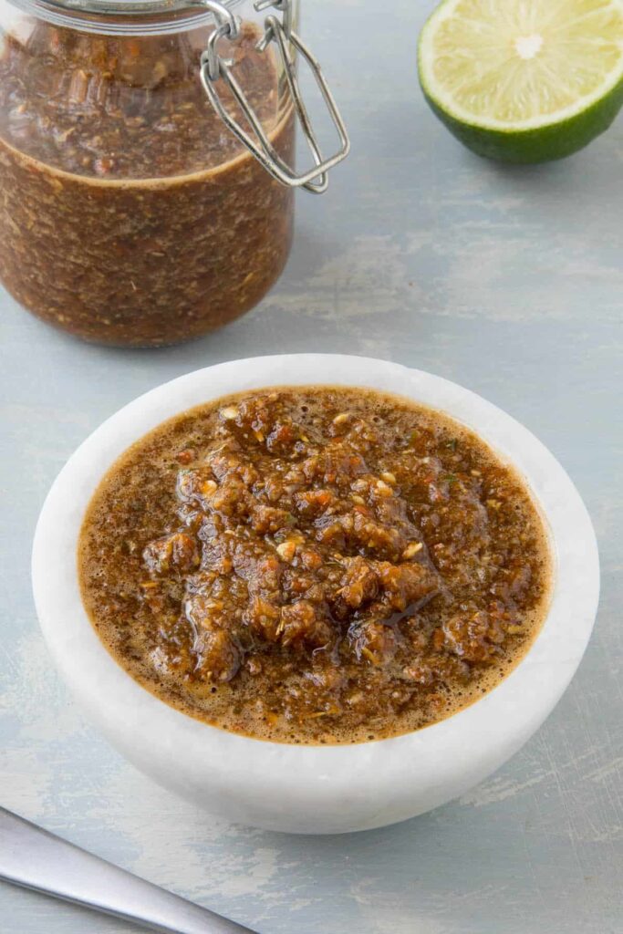 A glass container of Jamaican jerk sauce with a small portion poured into a bowl with half a lime sitting next to it. 