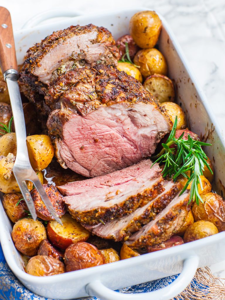 Baking dish filled with a leg of lamb, potatoes and some fresh rosemary. 