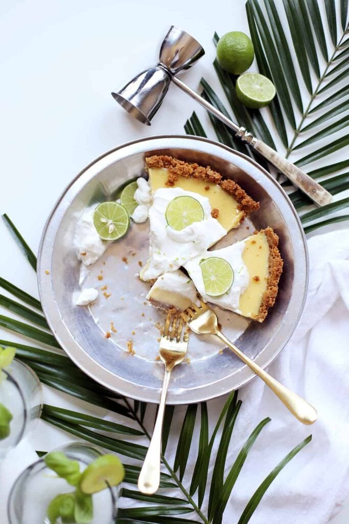 Te Key Lime Pie with most of the pieces gone, and 2 forks next to the remaining pieces and fresh limes lying next to the pie plate. 