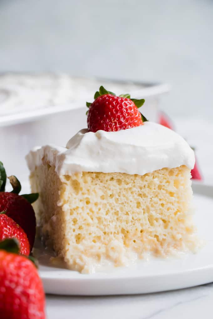 A few fresh strawberries next to a piece of tres leches cake, topped with fresh whipped cream. 