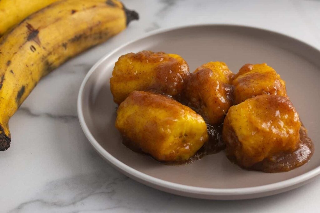 Caramelized plantains on a plate with an unpeeled plantain next to it. 