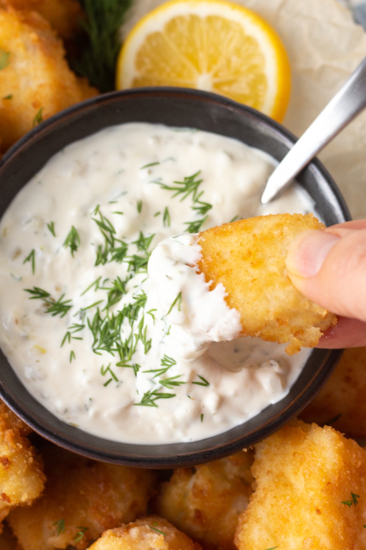 Small bowl of tartar sauce with dill on top and a piece of fried walleye being dipped into it.