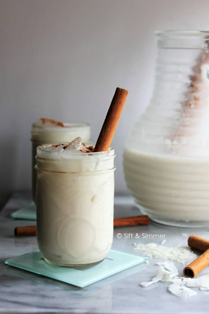 Horchata served in glasses with a cinnamon stick garnished in it in front of the pitcher of horchata. . 