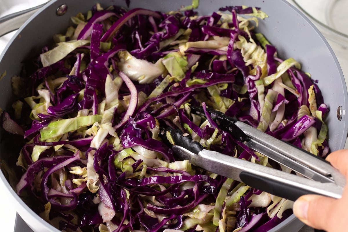Hand tossing cabbage in a skillet with a pair of tongs.