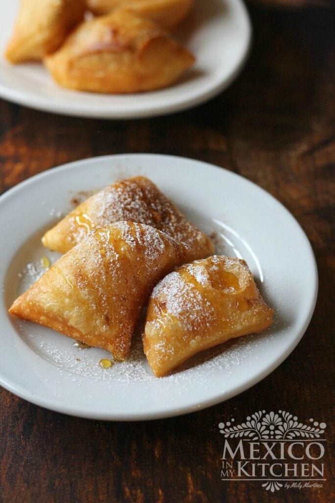 Sopapillas on a plate covered in powdered sugar and drizzled with honey. 
