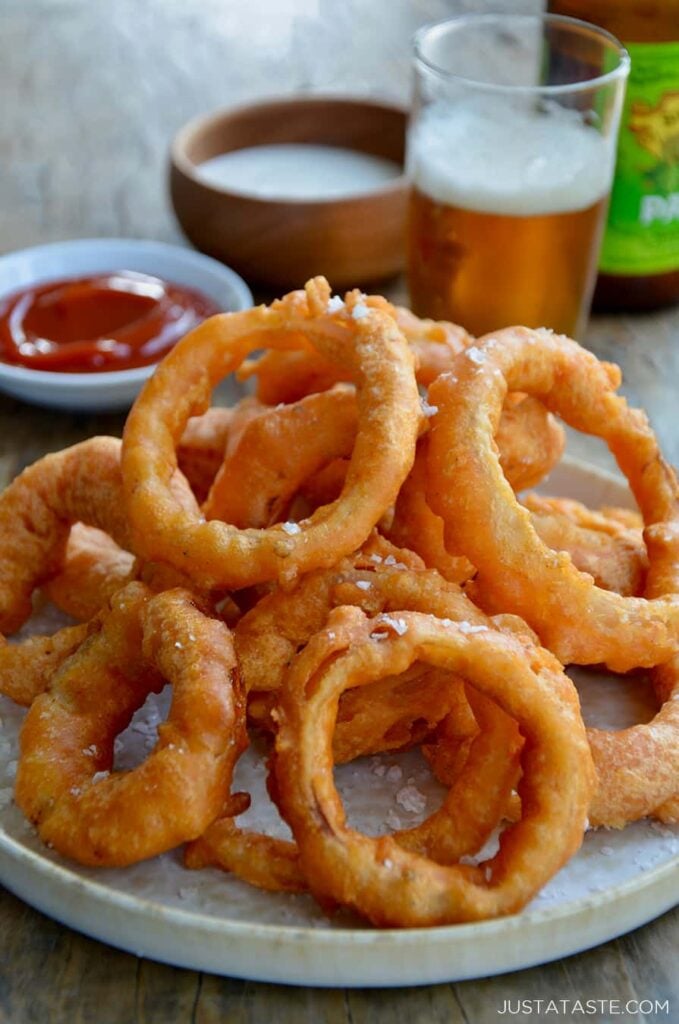 Beer-battered onioin rings piled on a plate with a side of ketchup and a glass of beer behind it. 