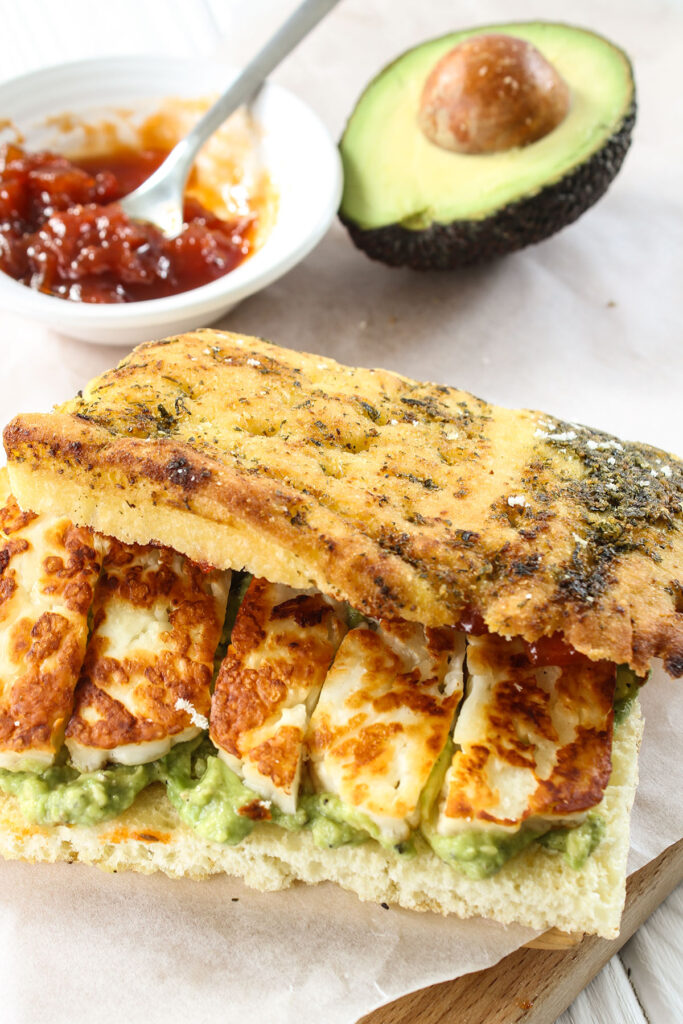 Halloumi and smached avocado sandwich with half an avocado and condiments sitting behind it. 