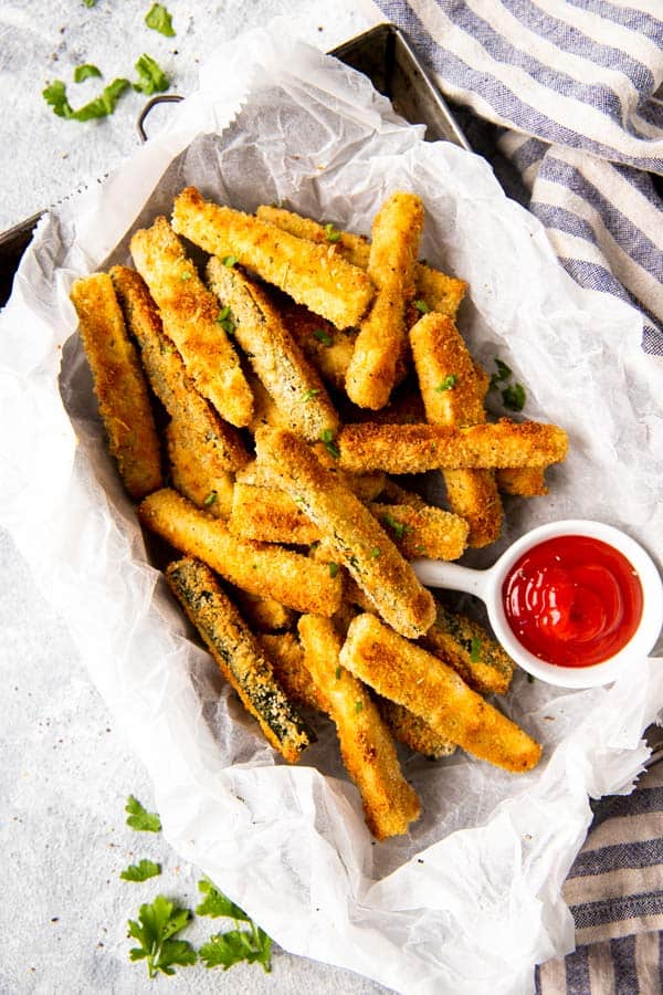 Parchment paper in a basket filled with zucchini fries and a small cup of ketchup. 