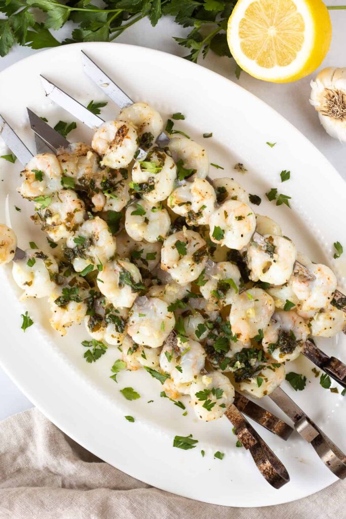 Metal skewers filled with grilled shrimp sitting on a plate with a halved lemon and garlic bulb sitting next to the plate. 