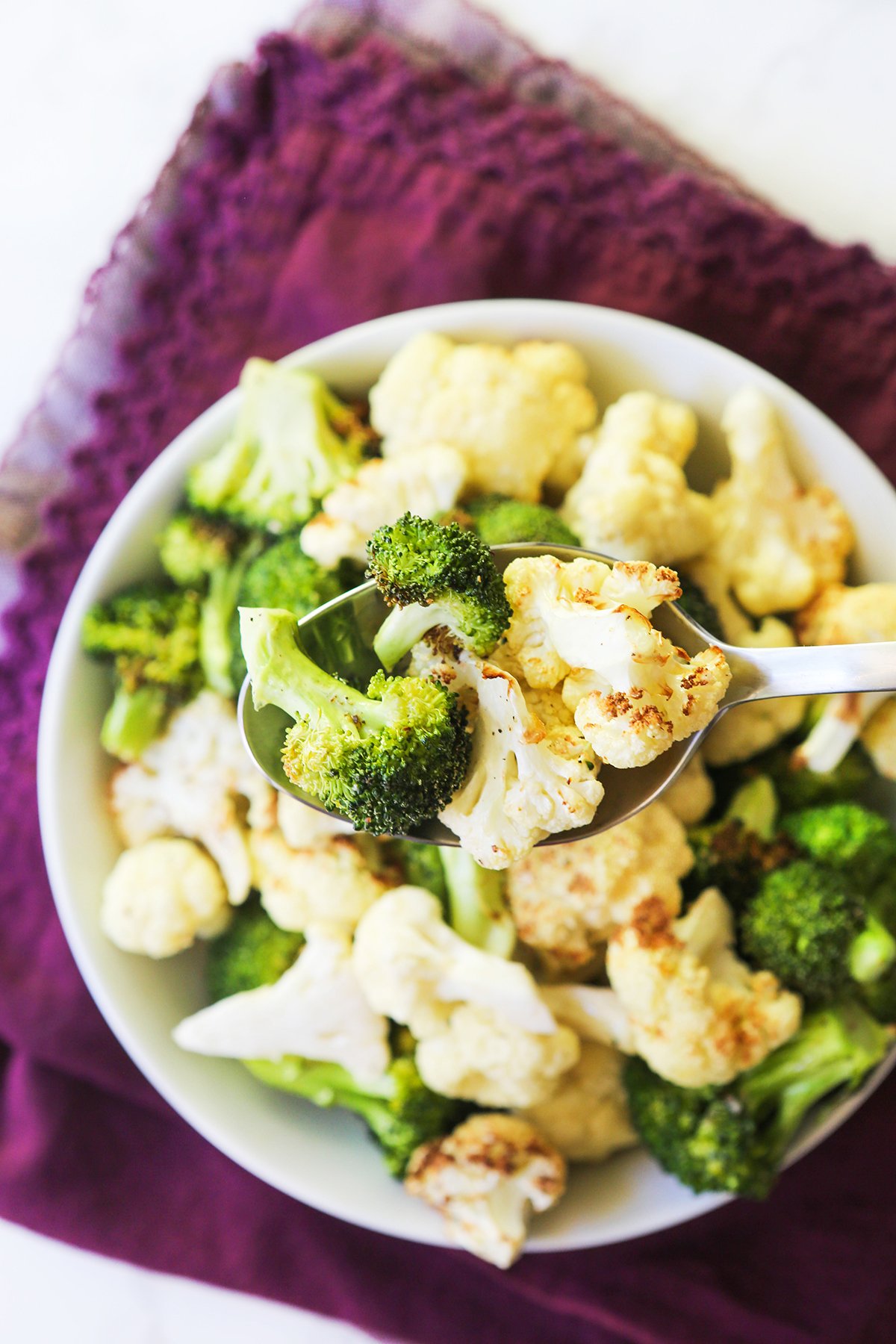 Spoon holding cooked broccoli and cauliflower florets over a serving bowl.