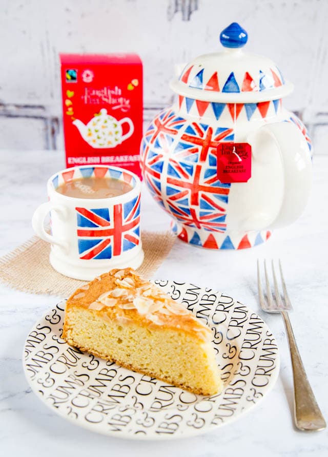 A tea pot and tea cup with a British flag on it filled with tea with a slice of cake on a plate. 