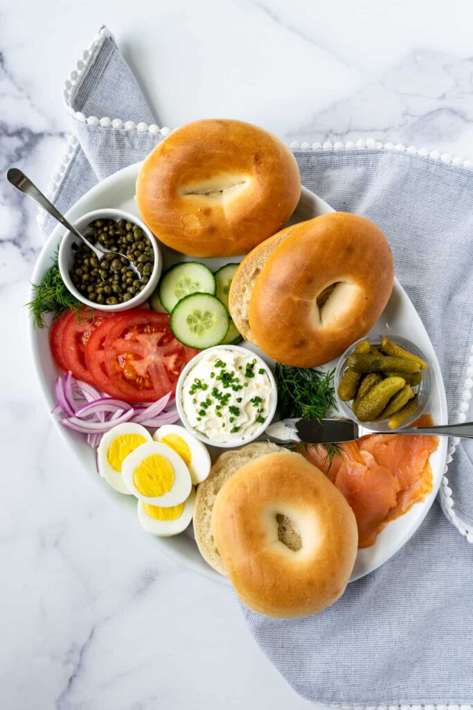 Smoked Salmon bagel platter with a variety of fresh veggies sliced, small bowl of baby dill pickles, a dig and whole bagels sliced in half. 