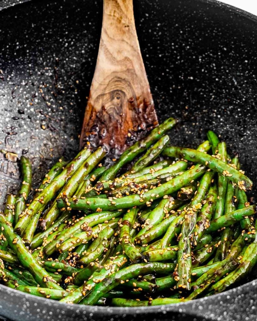 Sauteed green beans topped with sesame seeds in a cooking pan and a wooden spoon moving the beans around. 