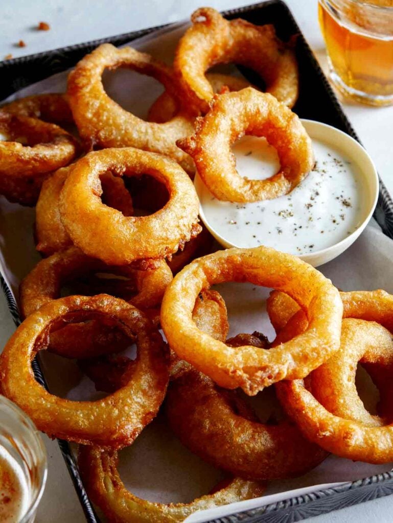 Beer battered onion rings on a platter with a small bowl of dipping sauce and one onion ring dipping into it. 