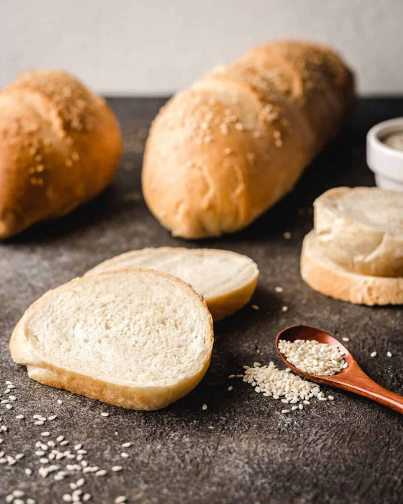 Loaves of semolina bread sitting behind slices of it with a spoonful of sesame seeds. 