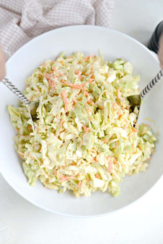 Classic coleslaw in a white serving bowl with metal salad tongs to serve with it. 