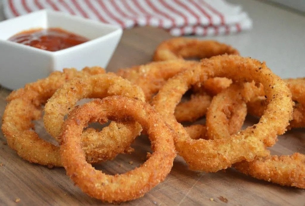 Extra crispy onion rings on a cutting board next to a small bowl of sauce. 