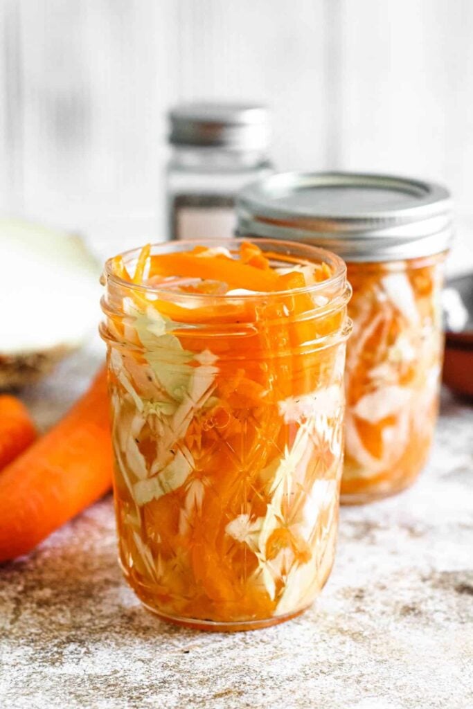 Two glass jars of pikliz with whole carrots laying next to it. 