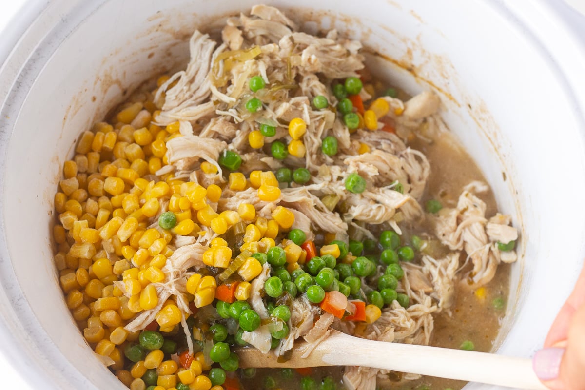Wooden spoon stirring corn, peas and shredded chicken in a crockpot.