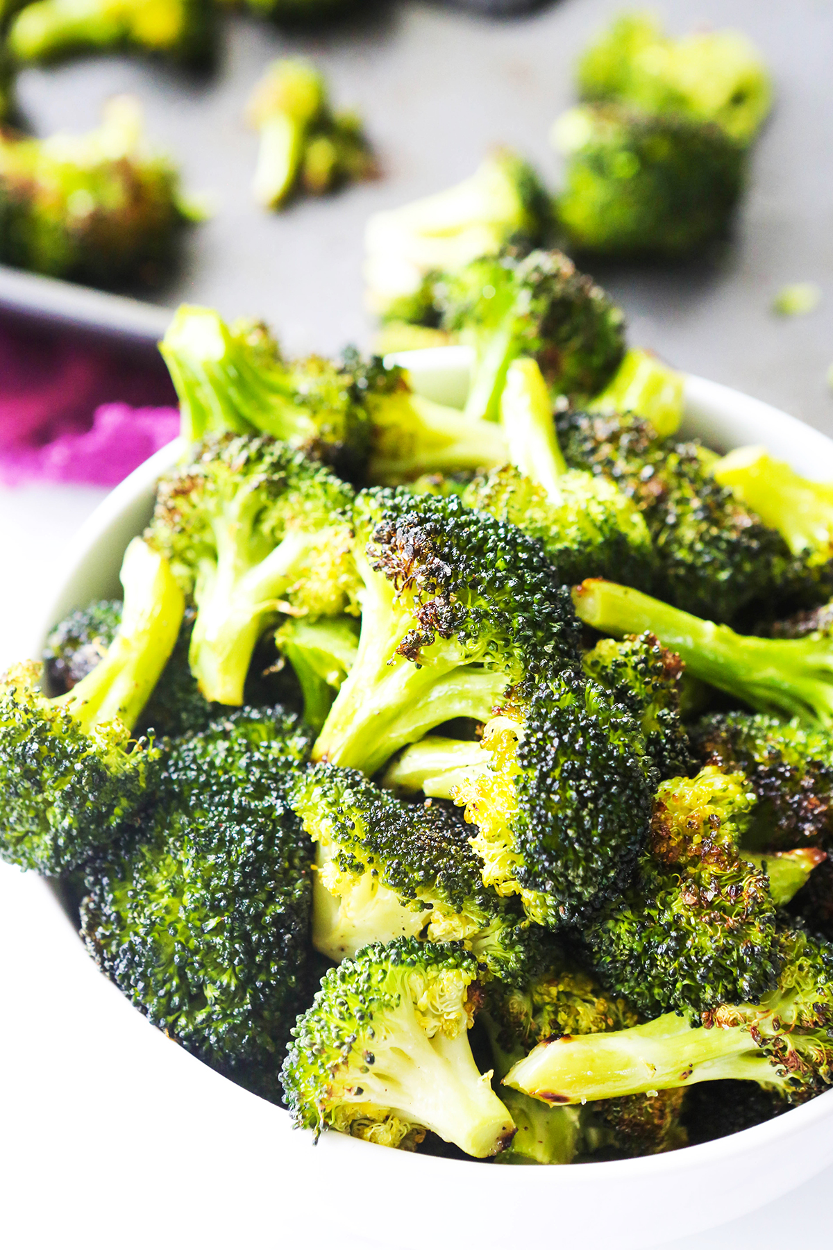 Roasted broccoli in a heaping pile in a white bowl, next to a baking sheet.