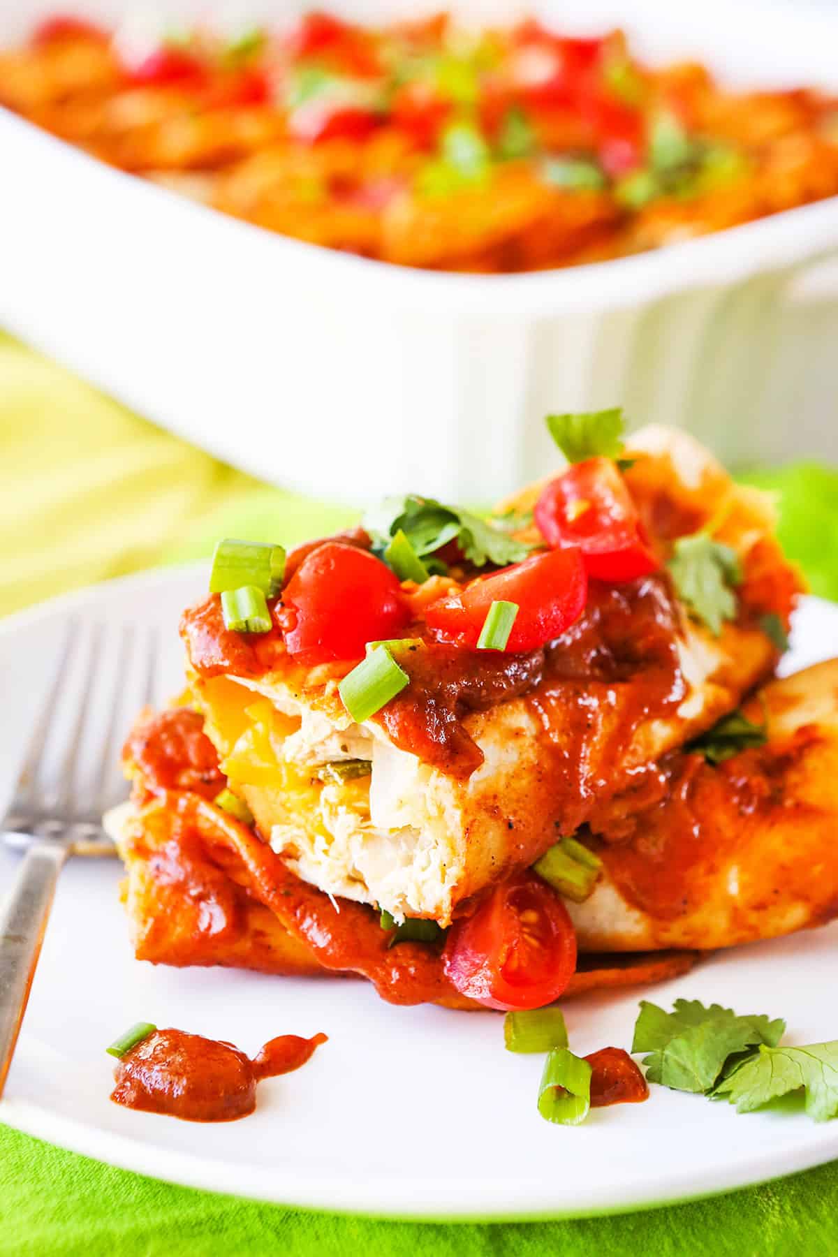 Stacked chicken enchiladas on a plate, sitting next to baking pan.