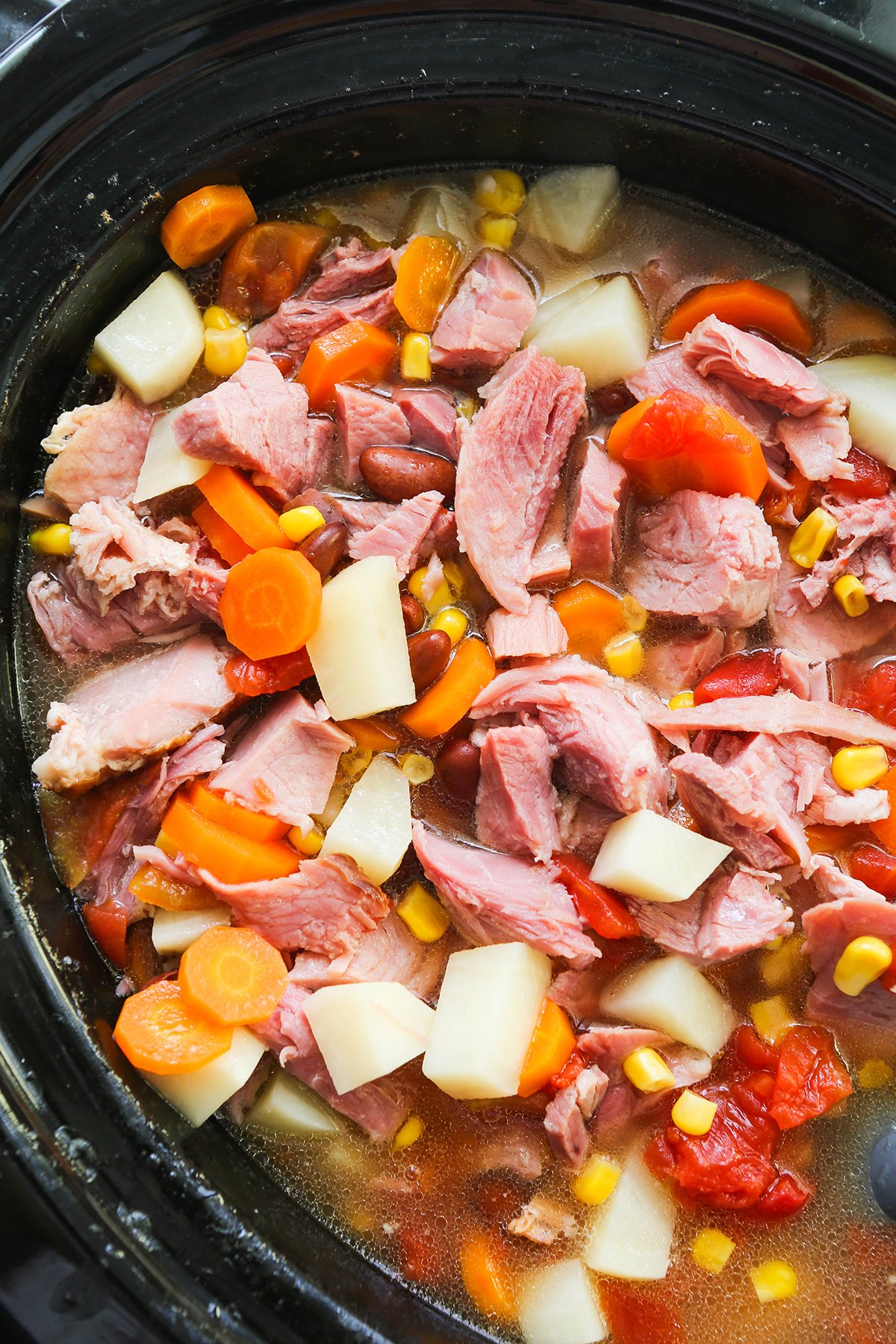 Ham pieces in a soup along with potato pieces and carrots, inside a slow cooker.