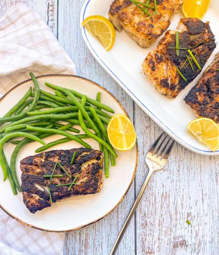 Blackened cod on a plate with green beans and a lemon wedge. 