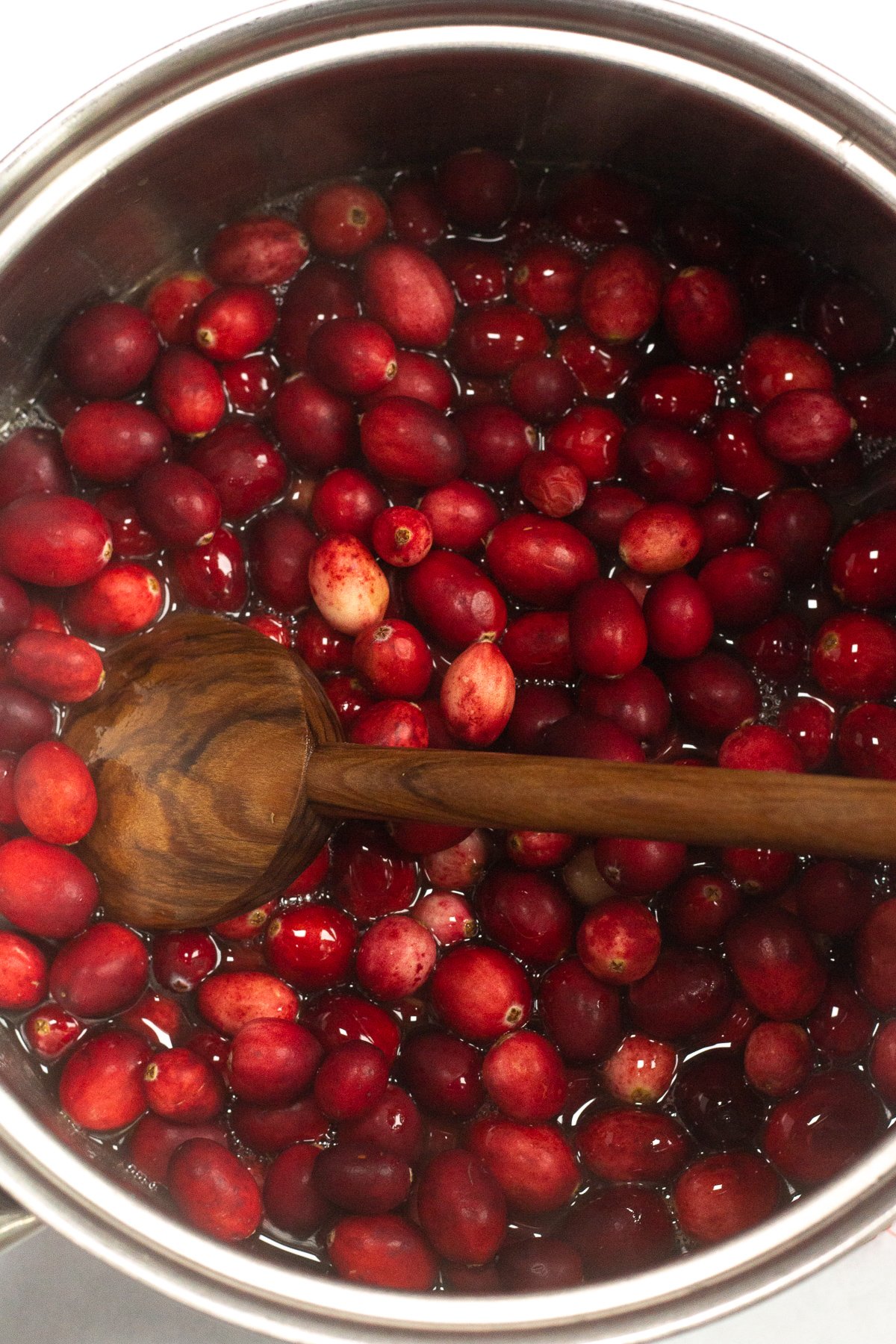 Fresh cranberries in a saucepan being stirred with a wooden spoon.
